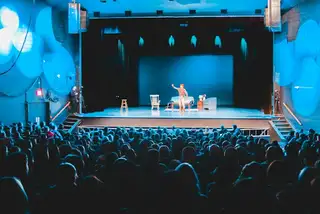 A crowded auditorium seen from the back row, the stage tiny and far away