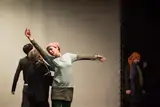 A performer in a headscarf reaching upward mid-movement while the cast moves around her