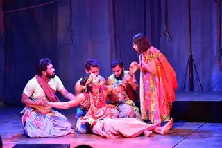A costumed cast gathered on a stage lit in blue and pink