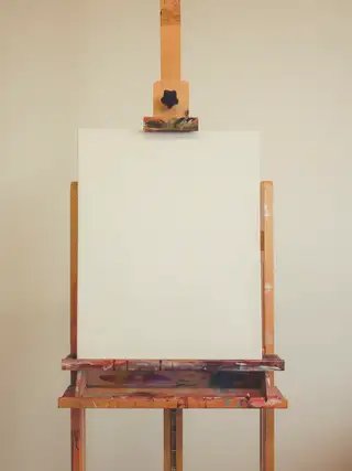 A wooden easel holding an untouched canvas in front of a cream wall