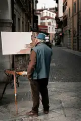 An older painter in a cap working at an easel on a narrow stone street