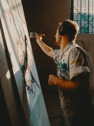 A painter in headphones and a paint-spattered shirt reaching up to work on a tall canvas