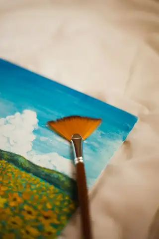 A small painting of a flower meadow under white clouds, with a fan brush resting across it