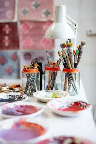 Jars of brushes and saucers of mixed pink paint on a white table by a lamp