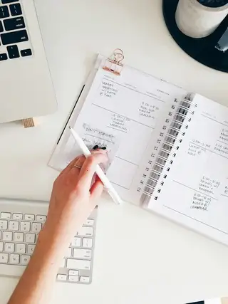 A hand writing in a spiral planner beside a laptop keyboard