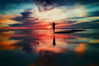 A figure with arms spread at the edge of a lake under a fiery sunset