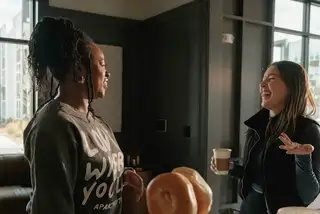 Two women laughing and talking in a sunny room, one holding a coffee cup