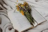 An open book with a handful of wildflowers lying on a soft white blanket