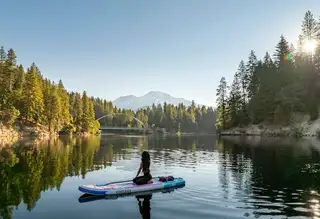 A person kneeling on a paddleboard on a calm lake below pine forest and a far mountain